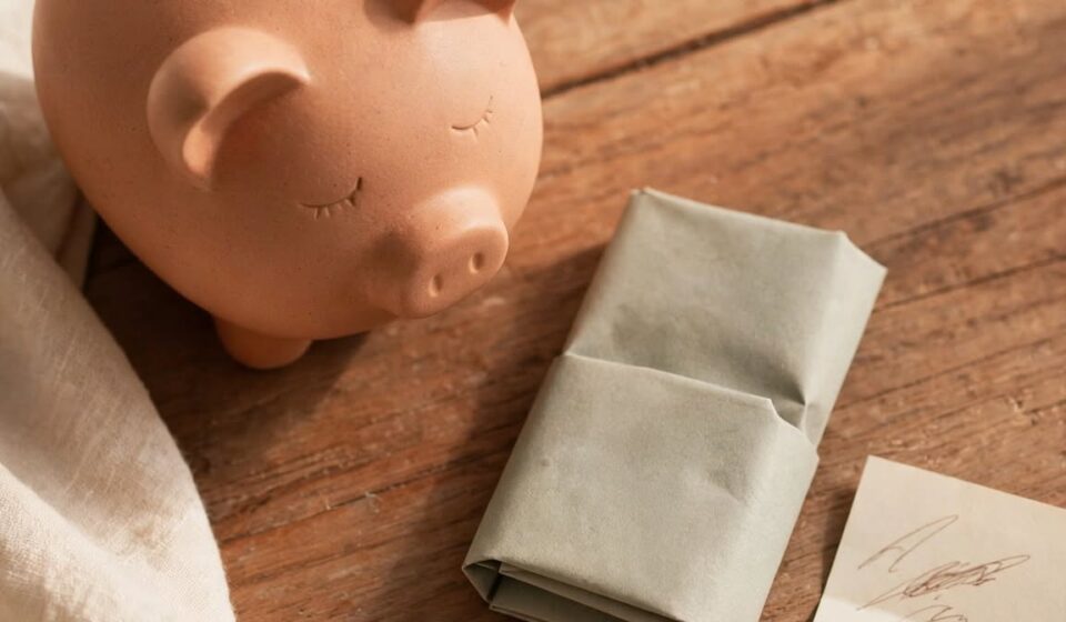 A piggy bank and folded bill on a wooden table, representing stewardship taught gradually before a teenager leaves home.