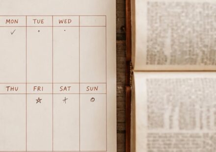 A weekly calendar with small marks spread across several days beside an open Bible, representing faith sustained throughout the week.