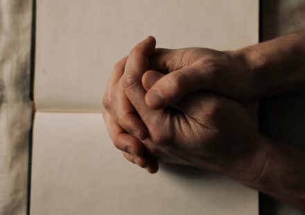 Clasped hands resting on an open Bible in dim evening light, representing honest prayer offered in a season of spiritual silence.