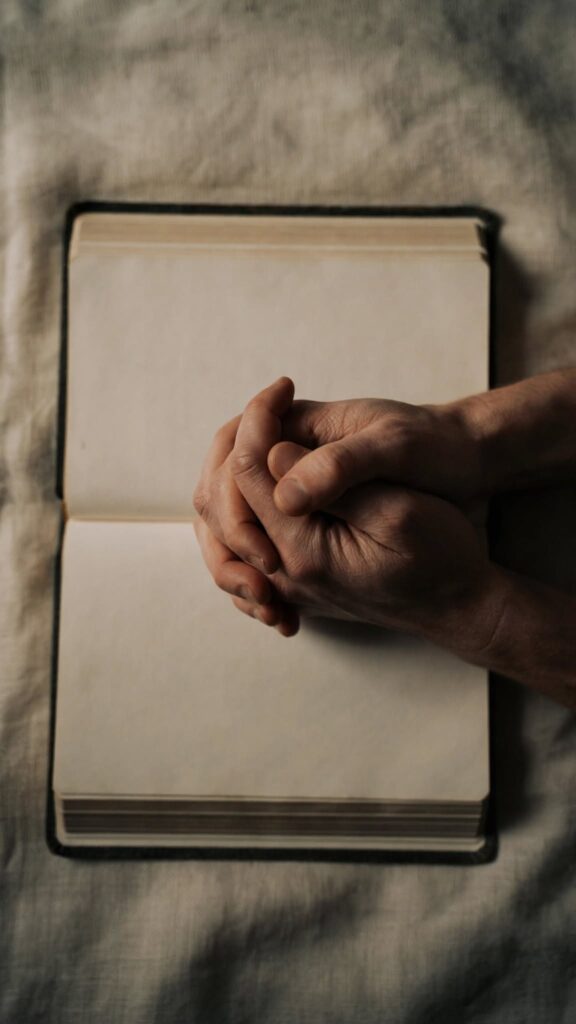 Clasped hands resting on an open Bible in dim evening light, representing honest prayer offered in a season of spiritual silence.