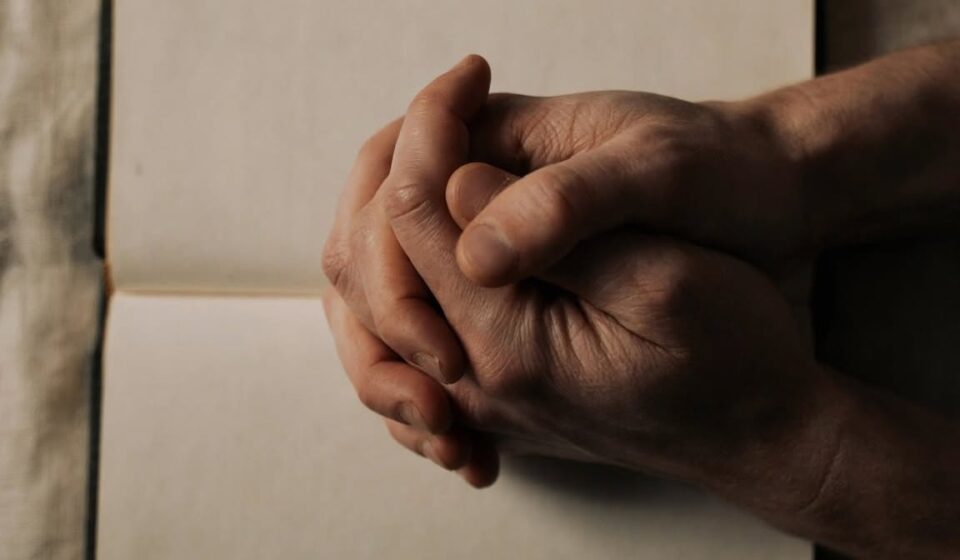 Clasped hands resting on an open Bible in dim evening light, representing honest prayer offered in a season of spiritual silence.