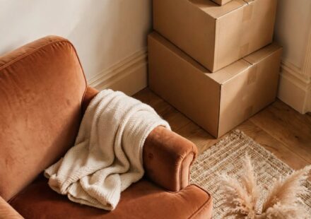 Moving boxes stacked beside a pushed-aside armchair in a cozy reading nook, representing a mother's space making room for her son's return.