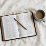 An open Bible and a lukewarm cup of coffee rest on cream linen in soft grey morning light.