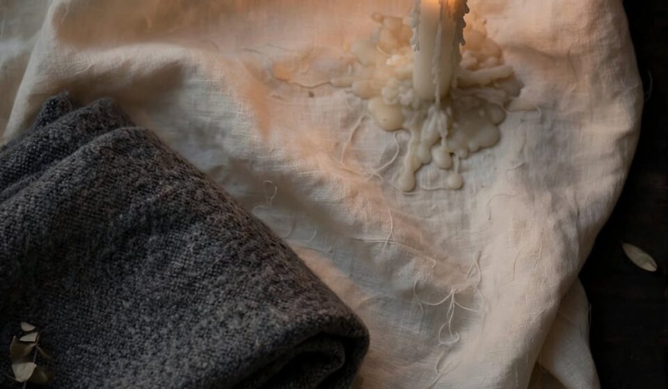 A single lit candle and a folded blanket sit on cream linen in soft dusk light.