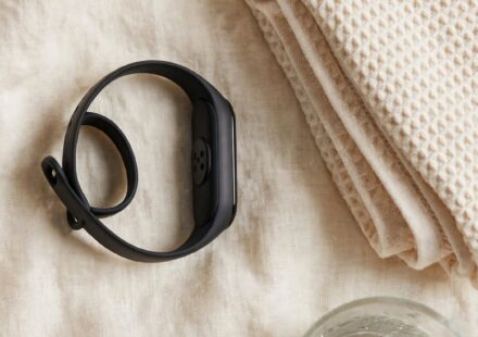 A fitness tracker rests face-down beside a folded towel and a glass of water on cream linen in soft natural light.