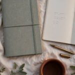 How to Do a Year-End Review as a Christian Before You Set a Single Goal A closed new planner sits beside an open notebook, a pen, and a terracotta mug on cream linen in soft evening light.