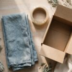 How to Sort Through a Parent’s Belongings Without Rushing Your Grief A folded blue tea towel, a roll of packing tape, and an open cardboard box sit on a wooden counter in soft morning light.