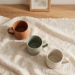 Living With Roommates as an Adult and Still Having a Real Home Three mismatched mugs sit on a wooden table beside a small framed picture leaning against the wall in soft morning light.