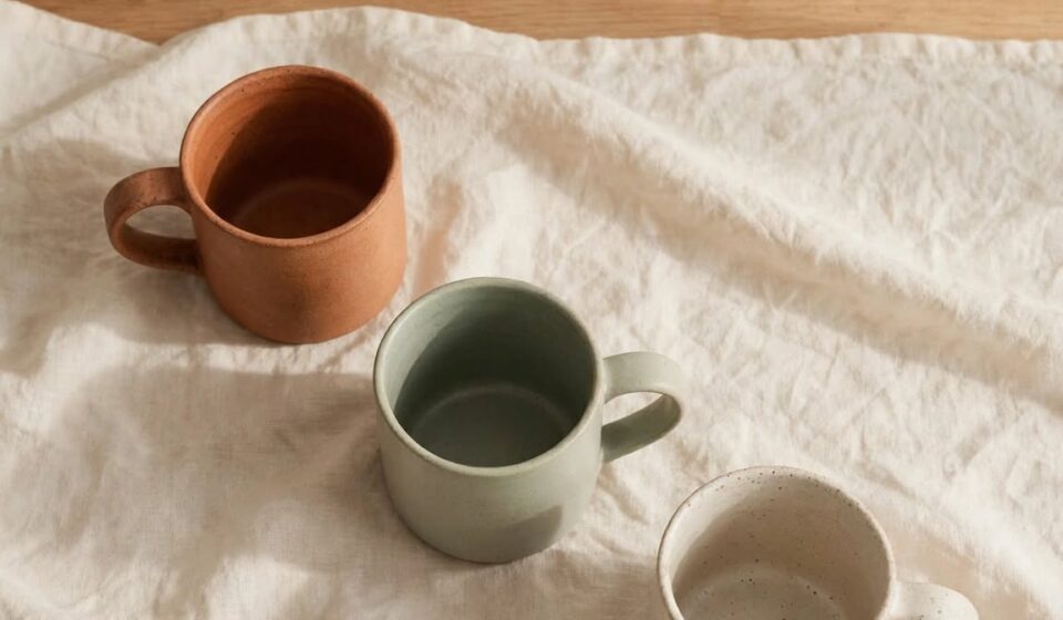 Three mismatched mugs sit on a wooden table beside a small framed picture leaning against the wall in soft morning light.