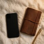 A phone lies face-down beside a closed journal and pen on cream linen in soft evening light.